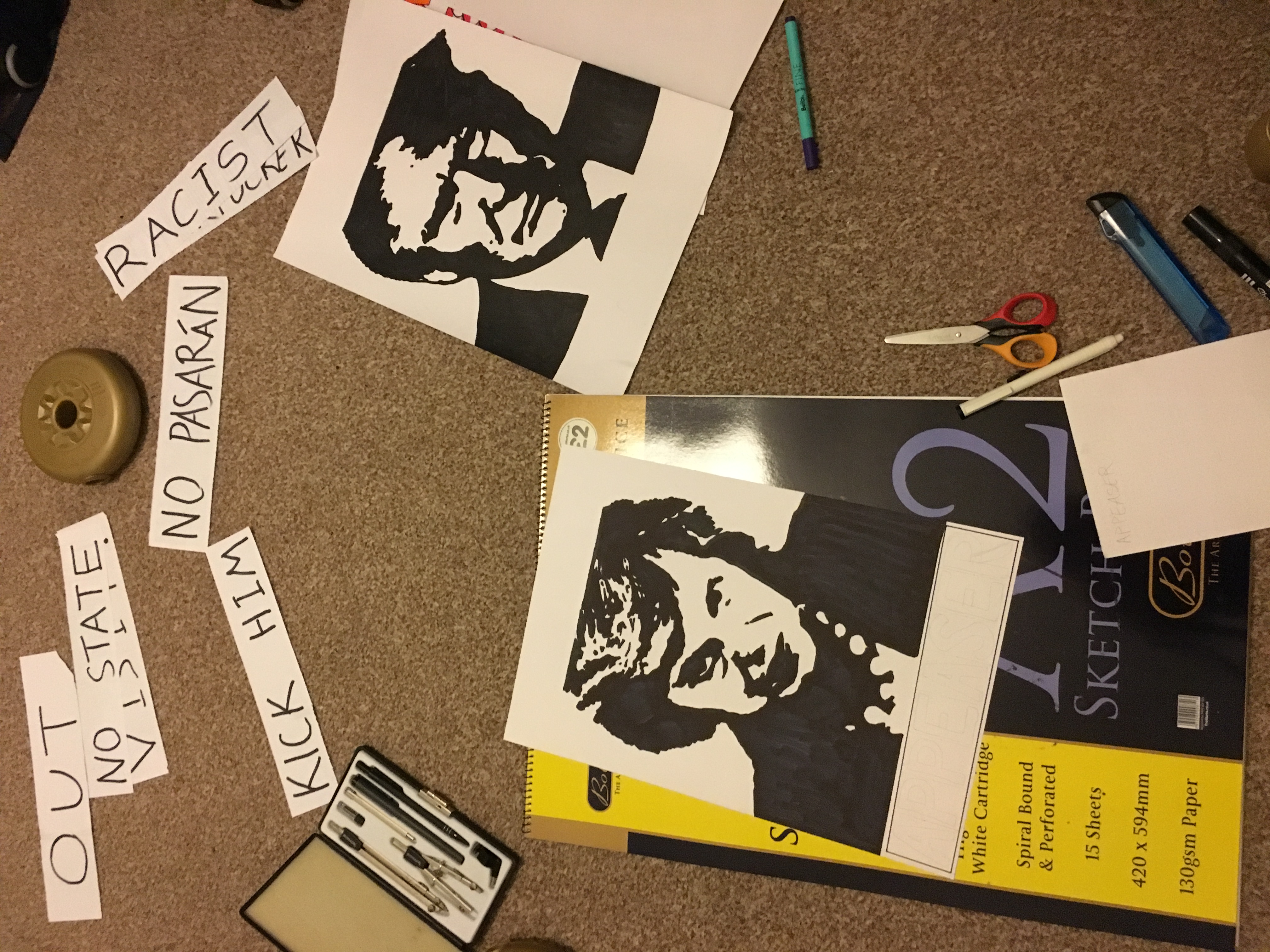 Placards showing stencil-style portaits of Theresa May and Donald Trump, in process of being made. A few slogans around on bits of paper "KICK HIM", "NO PASARÁN", "RACIST", etc.